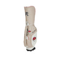 Beige Malbon Golf cart bag with wheels featuring elegant script logo, red trim, and cartoon mascot patch. High-quality leather-like material with multiple pockets and durable construction for professional golfers.