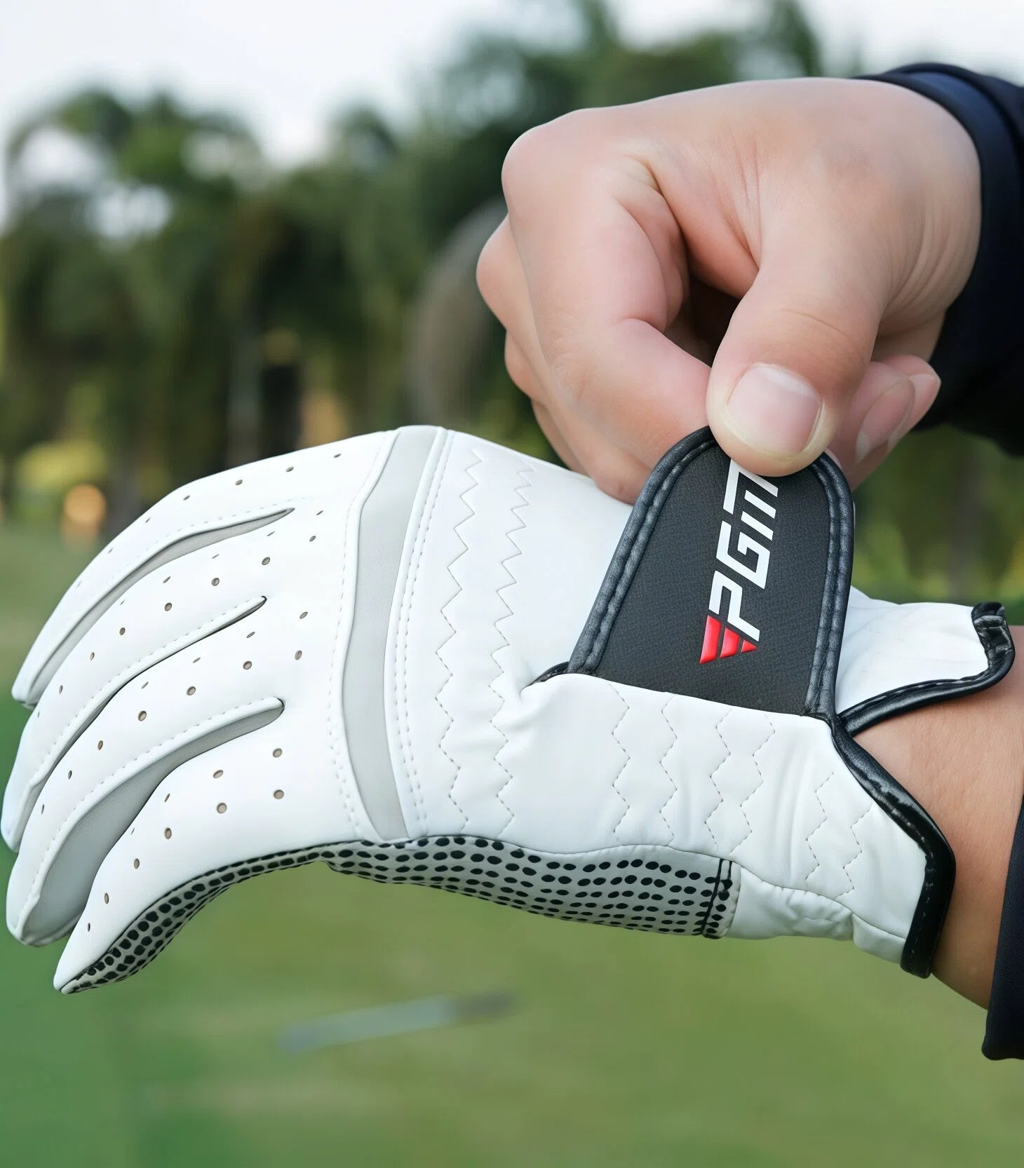 A hand adjusts the black Velcro wrist strap on the white PGM leather golf glove. This feature ensures a snug and personalized fit for maximum comfort and stability during play.